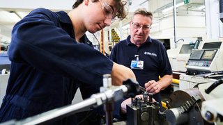 An engineering apprentice works, while his learning coach looks on.
