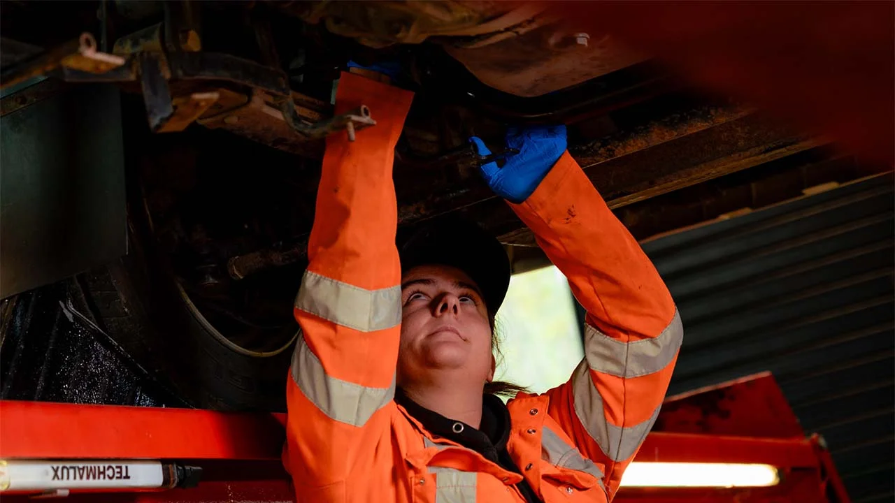An apprentice in an orange hi-viz jacket is working on the underside of a large vehicle that is raised off the ground.