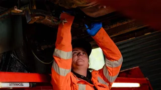 An apprentice in an orange hi-viz jacket is working on the underside of a large vehicle that is raised off the ground.