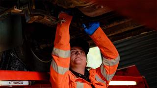 An apprentice in an orange hi-viz jacket is working on the underside of a large vehicle that is raised off the ground.