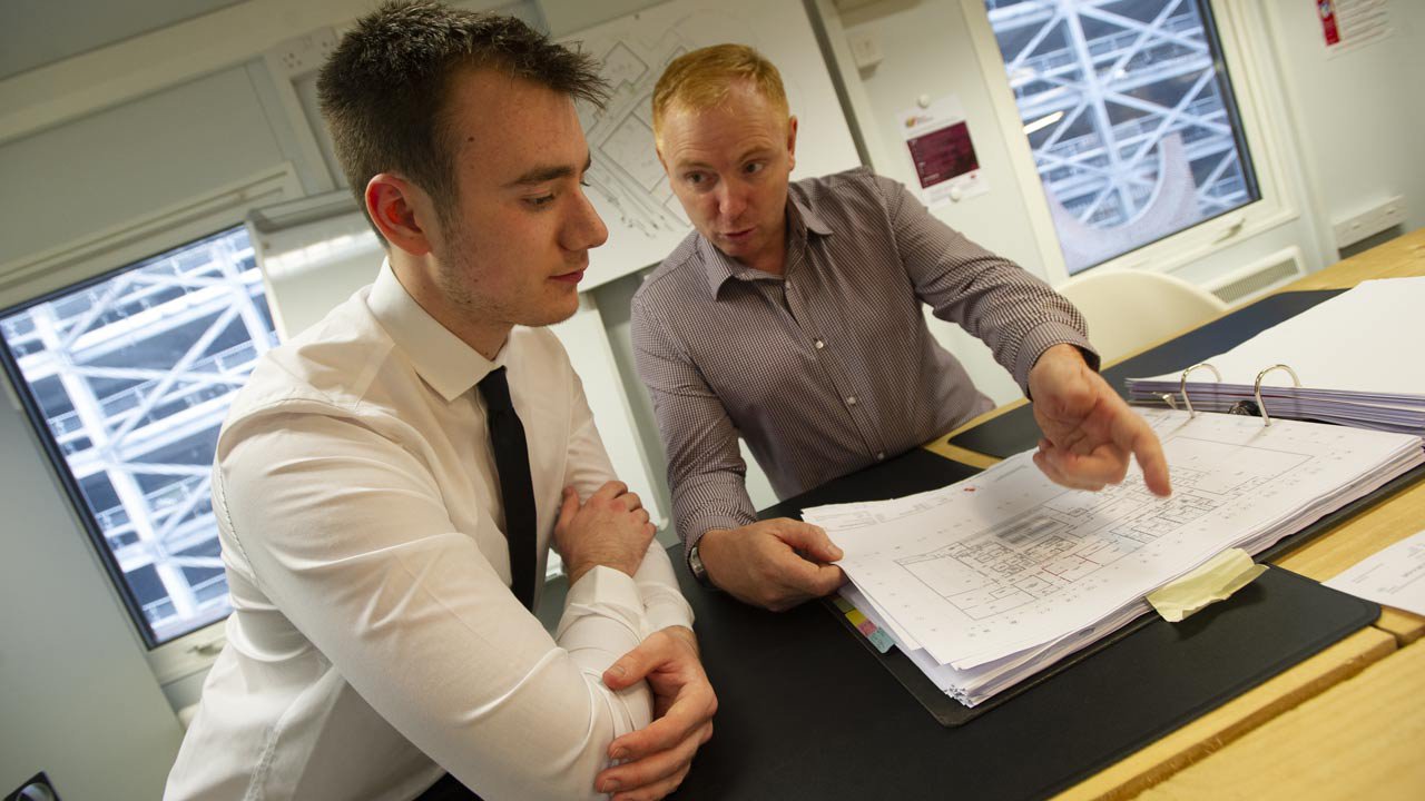 An apprentice and their manager discuss building plans in an office.