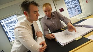 An apprentice and their manager discuss building plans in an office.