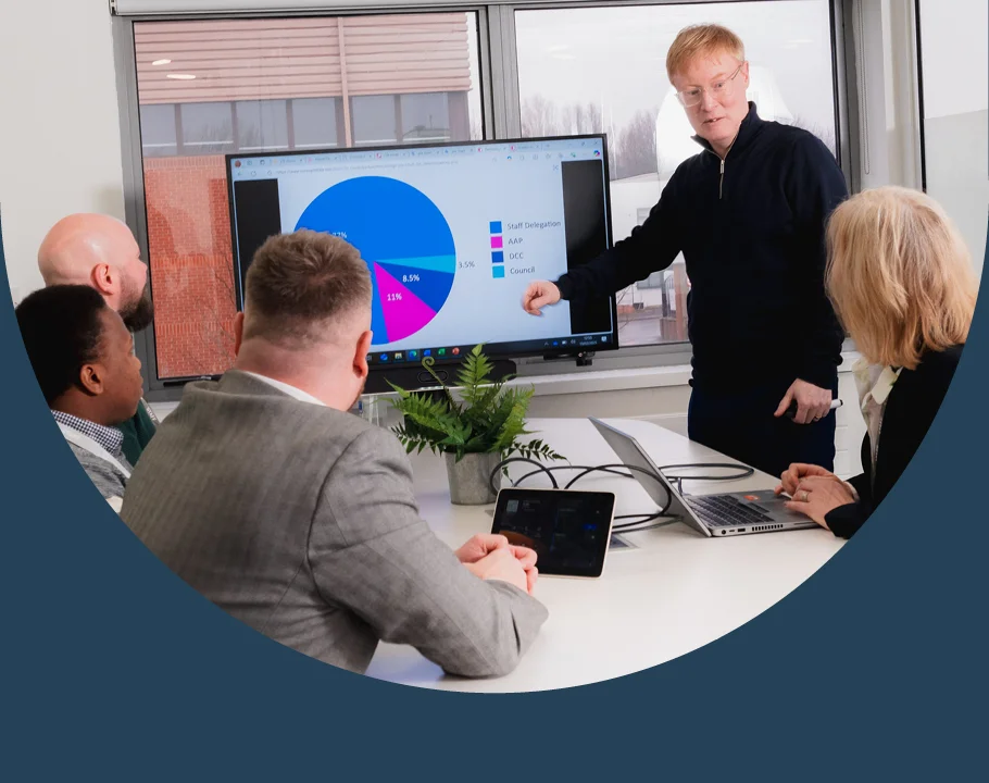 People attending a meeting while a presenter points to a pie chart displayed on a screen in a conference room.