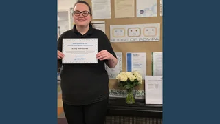Kelly Ann Jones is smiling at the camera with a certificate for her Personal Development Award in hand. She's standing in front of a wall display that says 'The House of Rompa', the name of the nursery,