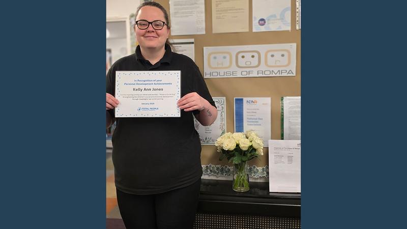 Kelly Ann Jones is smiling at the camera with a certificate for her Personal Development Award in hand. She's standing in front of a wall display that says 'The House of Rompa', the name of the nursery,