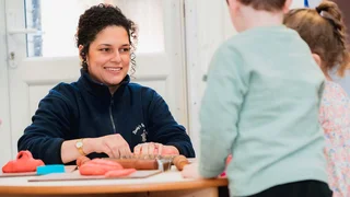 An Early Years apprentice is in a nursery setting, across a small table from two young children.