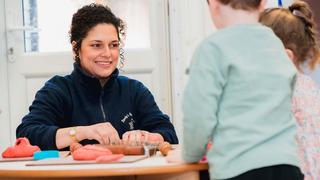 An Early Years apprentice is in a nursery setting, across a small table from two young children.