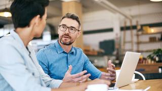 Two male colleagues are having a discussion while sat at a desk in their office, with a laptop open.