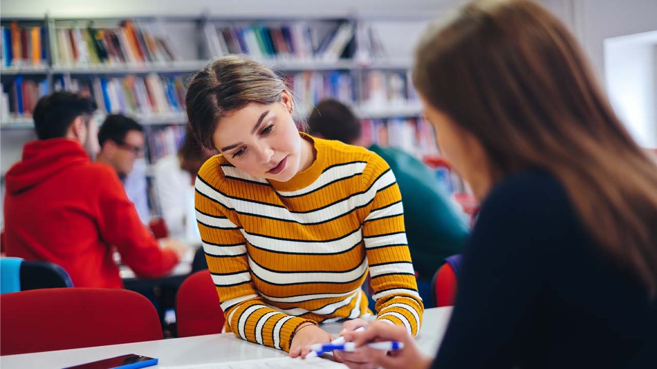 Two young women are sat at opposite sides of a desk in a library setting while going through an open book on the table together.