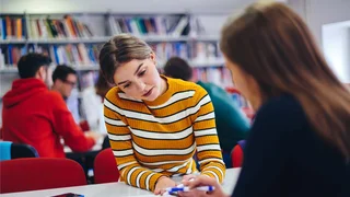 Two young women are sat at opposite sides of a desk in a library setting while going through an open book on the table together.