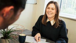A woman is sat across a desk from someone and smiling at them.