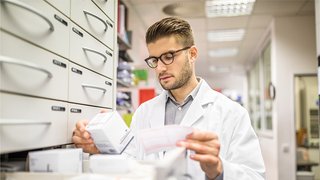 A pharmacist is picking out medicine from a drawer while checking a prescription.