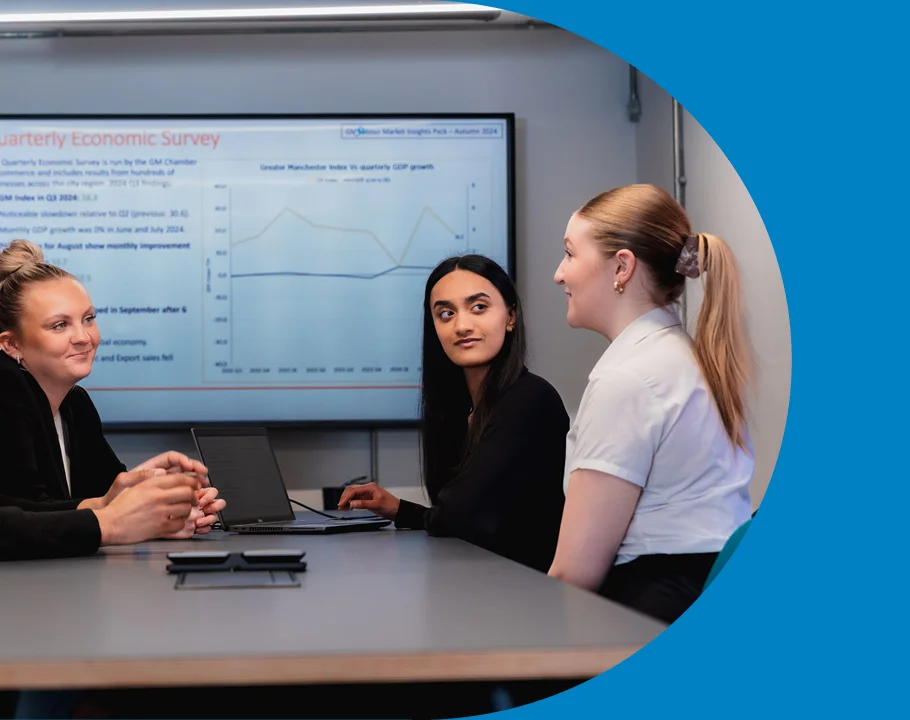 Three people in a meeting room discussing information displayed on a presentation screen, with a laptop on the table.
