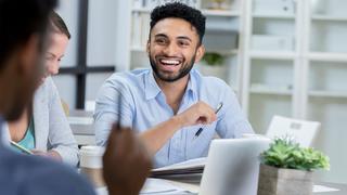 A man, the focal point of the picture, is laughing while working with his colleagues around a desk.