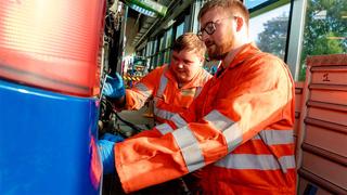 Two apprentices in orange high-vis clothing are working on a large vehicle in a workshop.