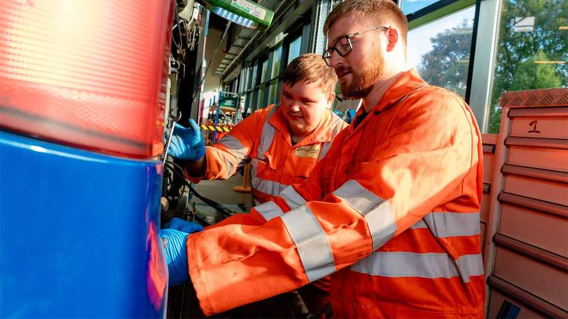 Two apprentices in orange high-vis clothing are working on a large vehicle in a workshop.