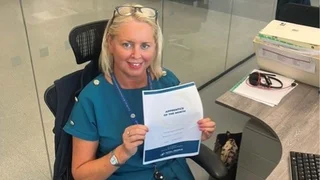 Rachel Lawson is sat at her office desk and holding her 'Apprentice of the Month' certificate while smiling at the camera.