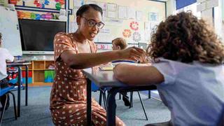A teacher kneels down at a desk to help a young pupil in a classroom.