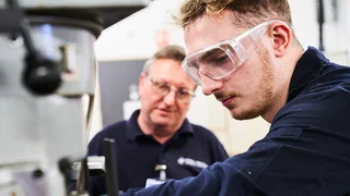 Apprentice working on engineering machinery with instructor guidance in a technical training workshop, wearing safety goggles and operating industrial equipment.