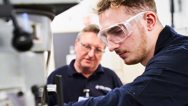 Apprentice working on engineering machinery with instructor guidance in a technical training workshop, wearing safety goggles and operating industrial equipment.