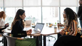 Two female colleagues are chatting in a bright, open office while colleagues work behind them.