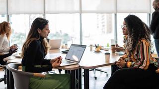 Two female colleagues are chatting in a bright, open office while colleagues work behind them.
