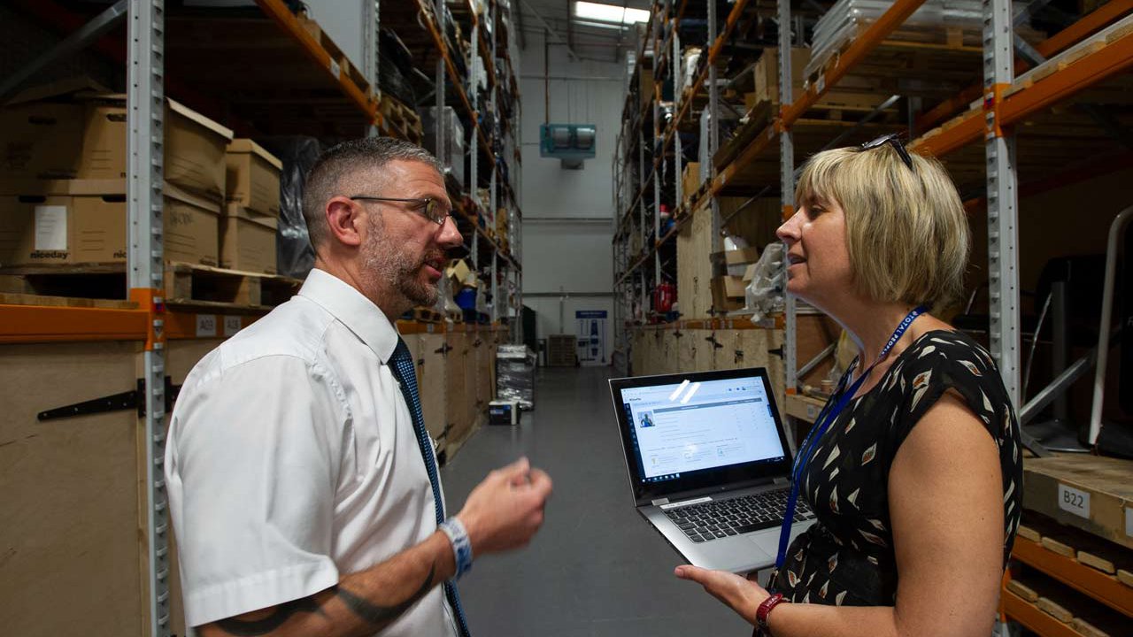 Man and woman holding a laptop speaking in a warehouse.