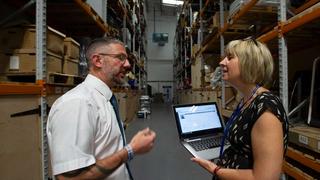 Man and woman holding a laptop speaking in a warehouse.