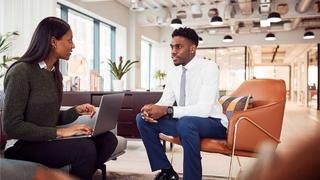 A young man chats to a woman in an open office setting as she types into a laptop.