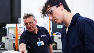 An Engineering apprentice in safety goggles is receiving instruction from a tutor in a workshop.