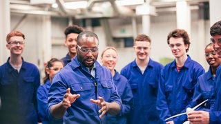 A group of students in blue overalls behind their teacher. Everyone is looking behind the lens at something the teacher is demonstrating.