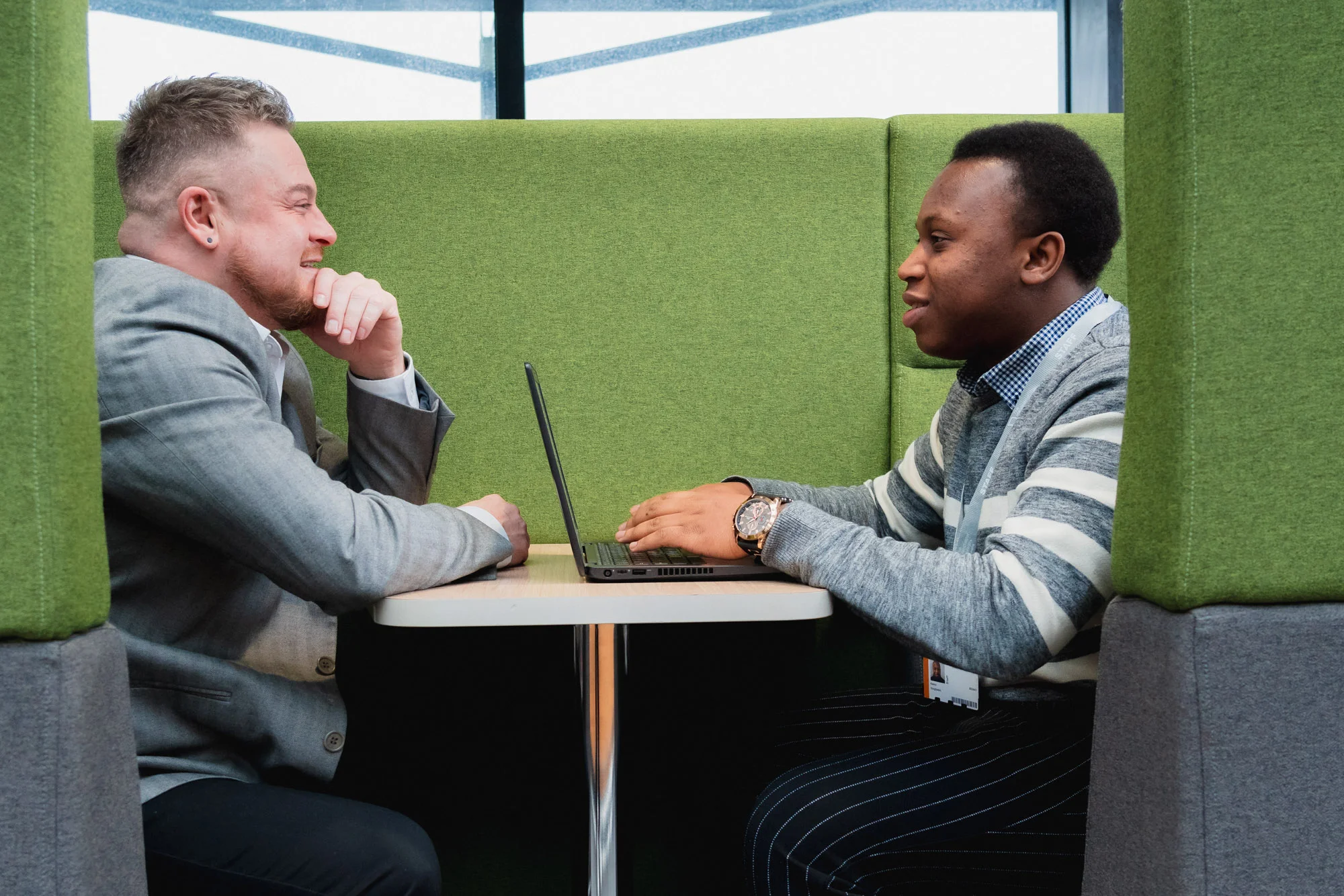 Two people sitting in a booth workspace discussing work across a table with a laptop.