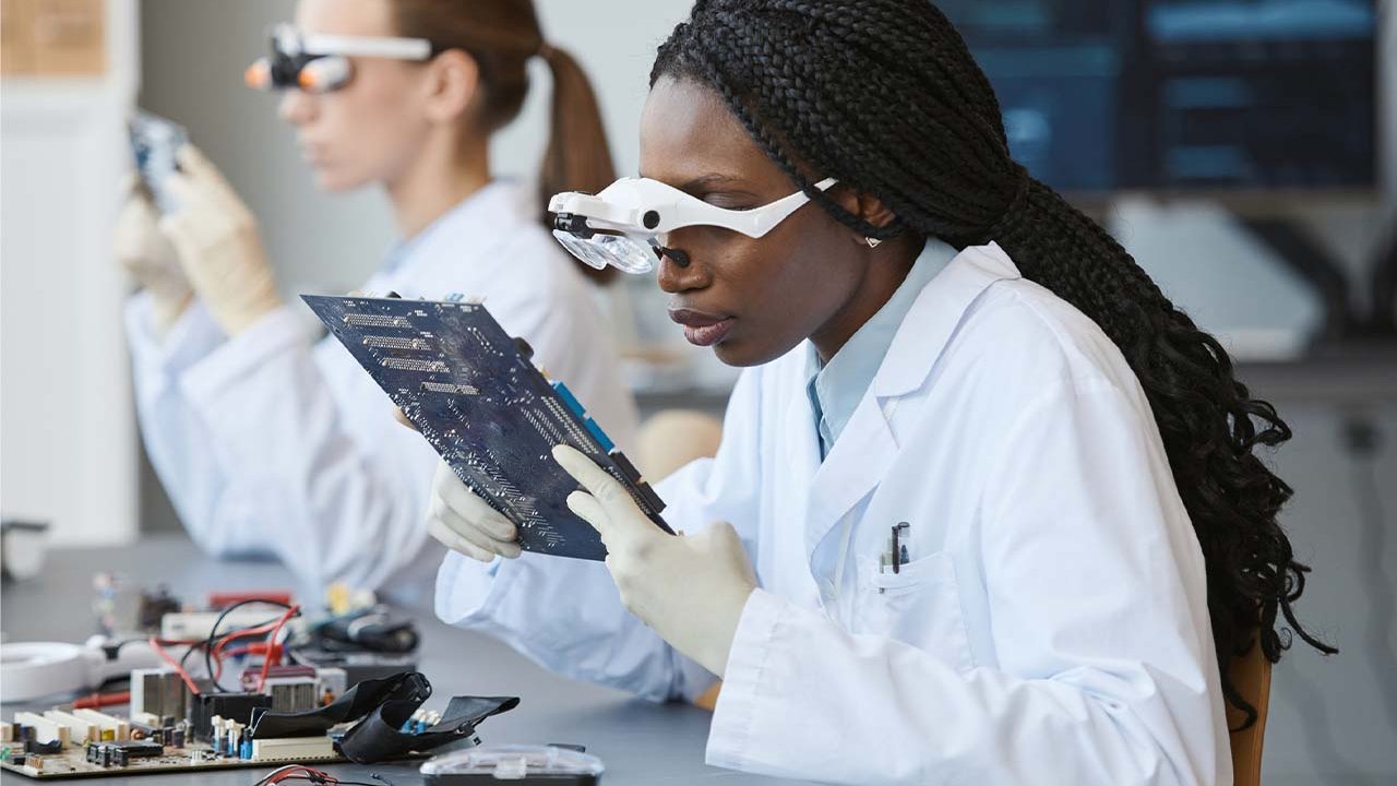 Two women in lab coats and goggles are at a work station inspecting pieces of equipment.