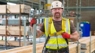 A man is standing on scaffolding within a large warehouse. He's wearing a hardhat and a high vis vest. He's looking directly at the camera and smiling.