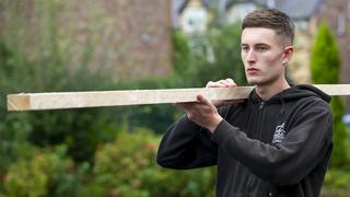 Male trades apprentice carries a plank of wood.