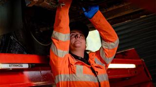 A young, female apprentice in orange overalls is working on the undercarriage of a large vehicle.