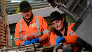 Two young apprentices are working under the bonnet of a bus in a workshop.