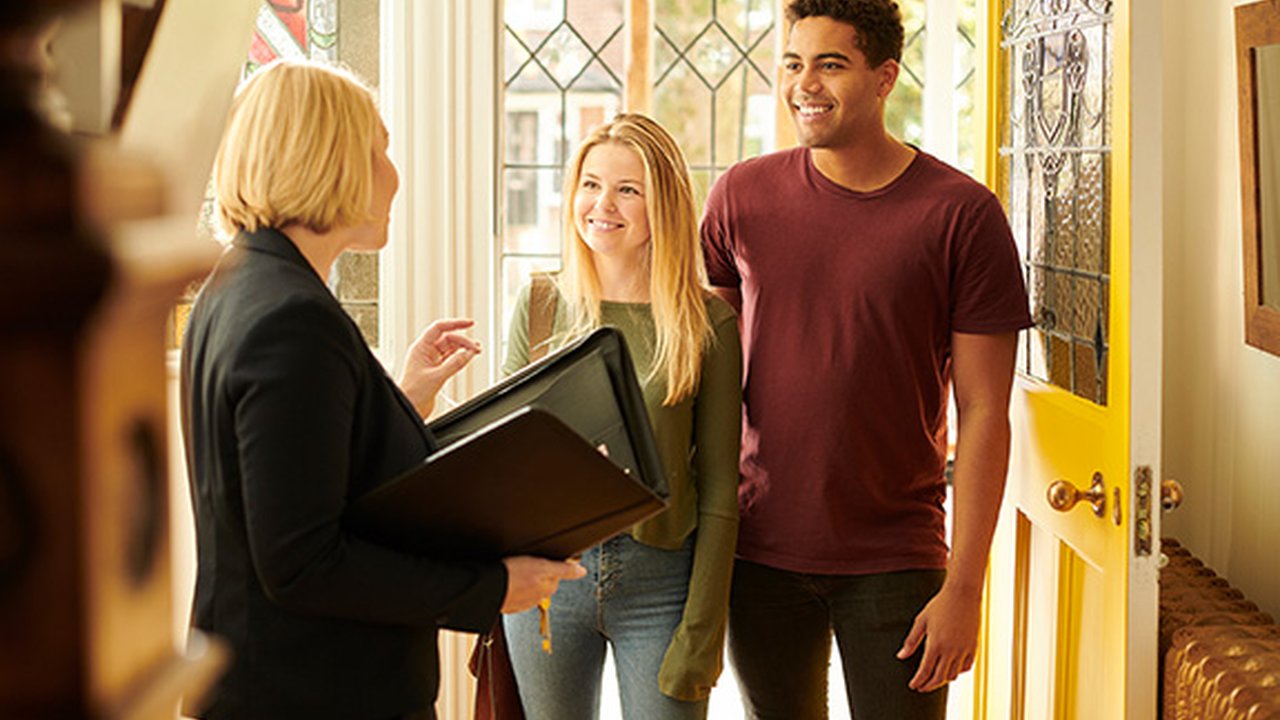 A couple are walking through the front door of a house as an estate agent welcomes them in.