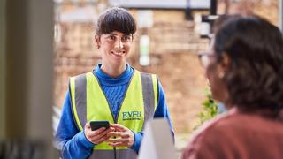 A delivery lady in an Evri uniform and hi-viz vest is stood at a front door delivering a parcel.