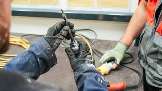 Close-up of two workers wearing protective gloves handling a drill bit and power tool attachment on a workbench.