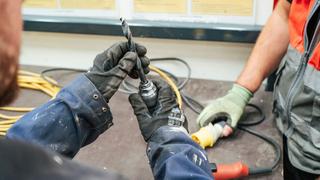 Close-up of two workers wearing protective gloves handling a drill bit and power tool attachment on a workbench.